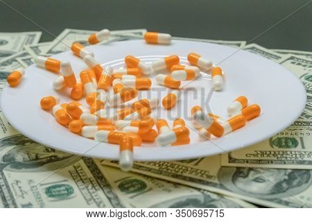 White Plate With Orange-and-white Tablets On The Table With Dollar Bills. Photos Of Falling Tablets 