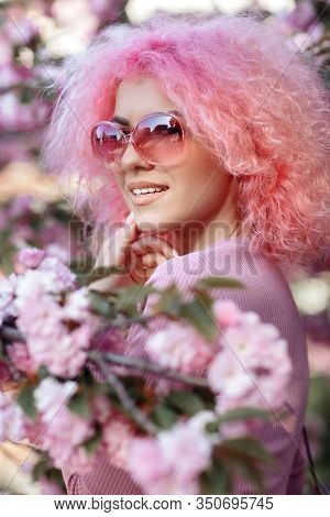 Close-up Portrait Of Attractive Young Woman With Curly Pink Hair And Sunglasses Near The Blossoming 