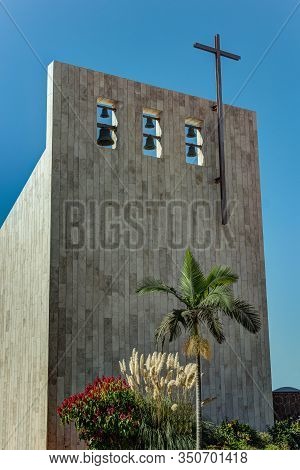 Cross On Church Over Clear Blue Sky. Six Bells Of Different Sizes On The Top. Beautiful Palm Tree An