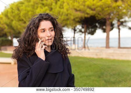 Thoughtful Woman Talking On Smartphone While Walking On Street. Serious Curly Lady Wearing Coat Comm