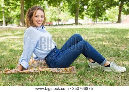 Beautiful Smiling Young Woman Sitting On Headkerchief In Park. Attractive Lady Wearing Casual Clothi