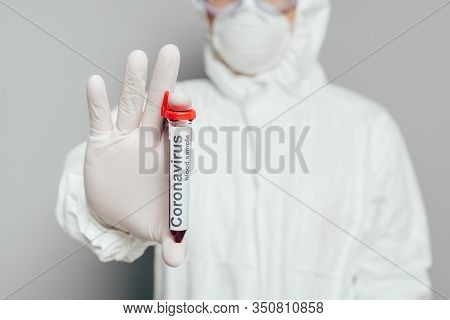Selective Focus Of Epidemiologist In Hazmat Suit Showing Test Tube With Blood Sample On Grey Backgro