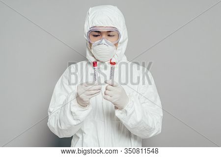 Asian Epidemiologist In Hazmat Suit And Respirator Mask Holding Test Tubes With Blood Samples On Gre