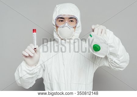 Asian Epidemiologist In Hazmat Suit Holding Respirator Mask And Test Tube With Blood Sample On Grey 