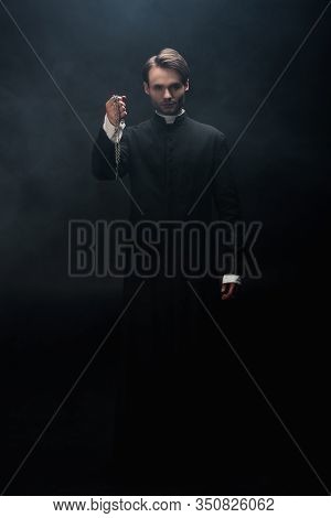 Full Length View Of Confident Catholic Priest Holding Necklace With Cross On Black Background With S
