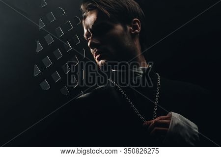 Young Serious Catholic Priest Touching Cross On His Necklace In Dark Near Confessional Grille