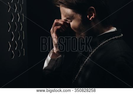 Young Tense Catholic Priest Touching Face With Closed Eyes Near Confessional Grille In Dark With Ray