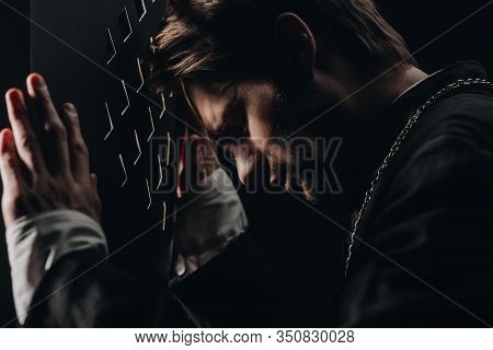 Young Tense Catholic Priest With Closed Eyes Leaning On Confessional Grille In Dark With Rays Of Lig