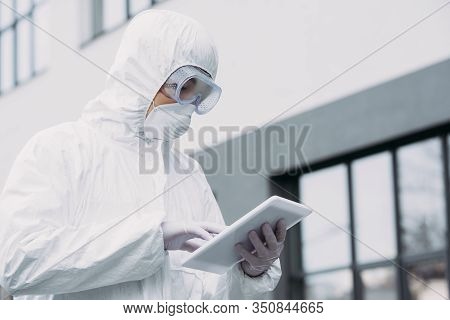 Asian Epidemiologist In Hazmat Suit And Respirator Mask Using Digital Tablet While Standing On Stree