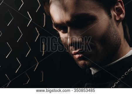 Portrait Of Young Tense Catholic Priest Looking At Camera Near Confessional Grille In Dark With Rays