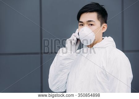 Asian Epidemiologist In Hazmat Suit And Respirator Mask Talking On Smartphone And Looking At Camera 