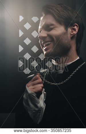 Sarcastic Catholic Priest Laughing While Touching Cross On His Necklace Near Confessional Grille In 