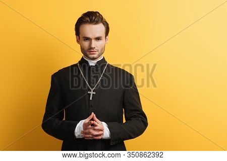 Serious Catholic Priest Standing With Clenched Hands While Looking At Camera Isolated On Yellow