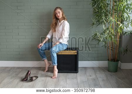 Young Beautiful Woman Sitting On Subwoofer With Bare Feet Next To Green Plant