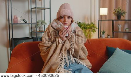 Young Sick Woman Wear Hat Wrapped In Plaid Sit Alone Shivering From Cold On Sofa Drinking Hot Tea In