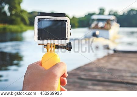 Action Camera Waterproof With A Yellow Float In A Man's Hand. In The Background Is A Wooden Pier Wit