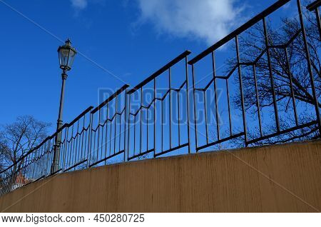 Paved Sidewalk With Gutter And Gray Metal Ornate Railings. Lamp With Glazed Lampshade And Black Post