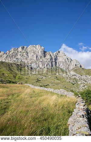 Green Meadows With Grass At The Foot Of A Rocky Mountain On A Sunny Summer Day, A Stone Wall Out Of 