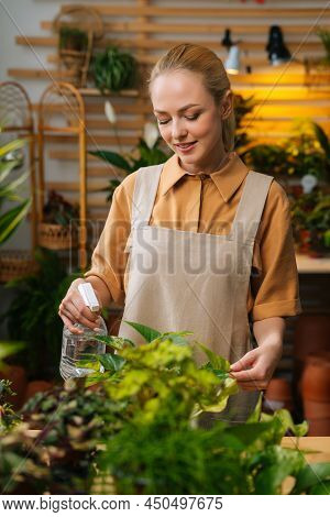 Vertical Portrait Of Positive Young Woman Florist In Apron Spraying Water On Houseplants In Flower P
