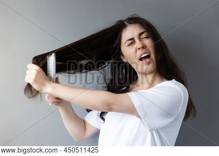 Portrait Of A Caucasian Young Woman Combing Her Tangled Dark Hair With An Effort With A Brush. Gray 