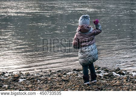 A Little Girl On A Winter Walk Stands With Her Back To Us And Throws Stones Into A Cold River