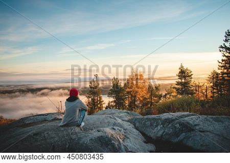 Hiker Sits Atop Vuokatinvaara In Sotkamo In The Kainuu Region, Finland At Sunrise And Above The Clou