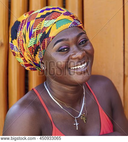 African Woman With Colorful Headdress And A Hopeful Happy Smile Stands Against A Painted Wooden Wall