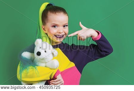 Little Model With Colored Hair Embracing And Pointing On Teddy Bear. The Concept Of Childrens Attach