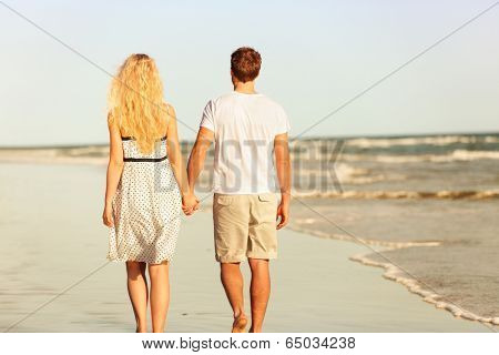 Beach couple holding hands walking at sunset by ocean sea. Beautiful casual young lovers hand in hand walking away with back at rear view. Summer vacation beach travel holidays concept.