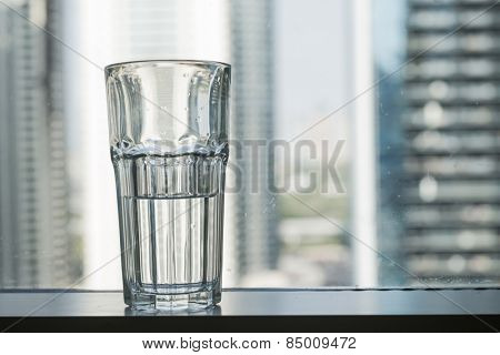 A half filled glass of water placed against window with skyline background. 