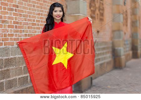 Pretty young Vietnamese woman with a lovely friendly smile holding up a the national flag of Vietnam as she stands in an urban street against a brick wall