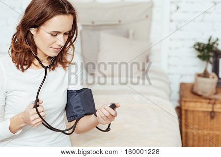 Under control. Pleasant charmign young woamn concentrating on measuring blood pressure and uisng tonometer while sitting on her bed