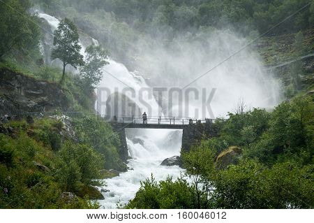 Wet tourist on the bridge and Briksdalsbreen big waterfall splashes. Norway popular touristic hike to Briksdal glacier.