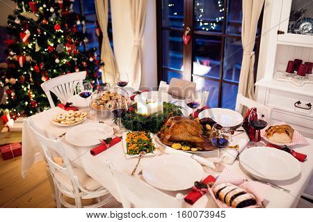 Christmas meal laid on table in decorated dining room. Roasted turkey or chicken, vegetables, cookies, Christmas tree, plates and glasses of red wine.
