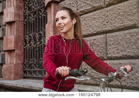Photo of young woman dressed in sweater walking with her bicycle near wall on the street and smiling while listening music. Look aside.