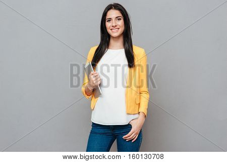 Young woman dressed in yellow jacket holding tablet computer over grey background. Look at camera.