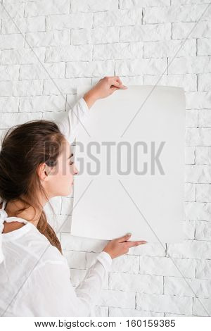 Woman holding empty whatman at wall mockup. Profile of young lady, looking at white sheet of paper, free space for advertisement or text. Warning message background