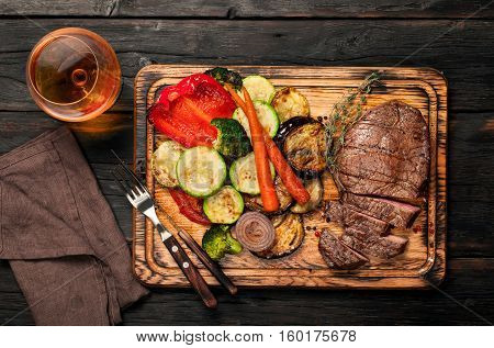 Slices of beef steak with grilled vegetables on cutting board on dark wooden table with a glass of brandy or whiskey