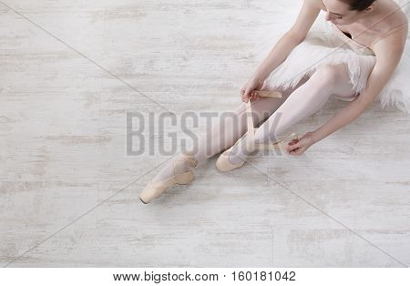 Beautiful legs of young ballerina who puts on pointe shoes at white wooden floor background, top view from above with copy space. Ballet practice. Beautiful slim graceful feet of ballet dancer.