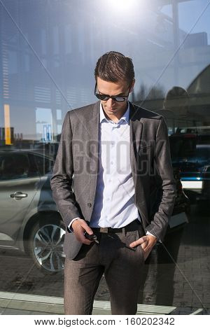 cars at the background, Male adult businessman in a suit  looking down and holding a car keys in his hand on a sunny cold day next to the car dealership building with big windows