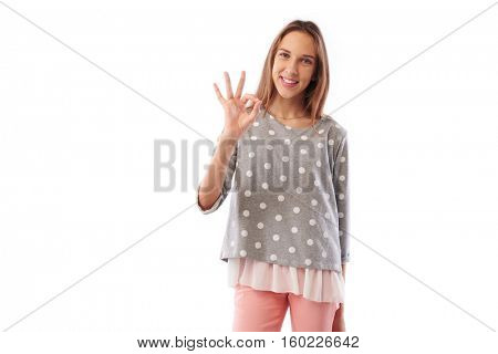 Mid shot of skinny happy toothy smiling young girl showing ok gesture standing over white background. Holding hand on hip while posing at the camera Concept of  nonverbal communication