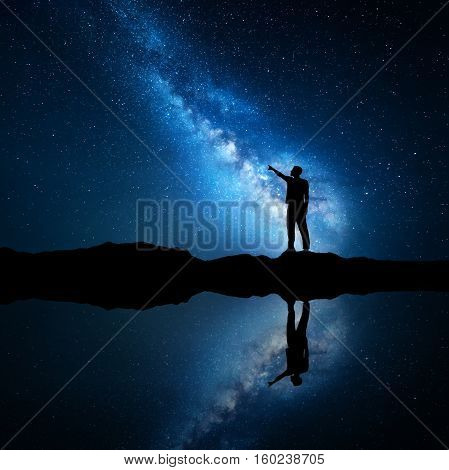 Milky Way. Silhouette of a standing man pointing finger in night starry sky on the mountain near the lake with sky reflection in water. Night landscape with universe. Blue milky way and man. Galaxy