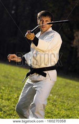 Kungfu expert in white kimono with black belt is practicing with nunchaku outdoors.