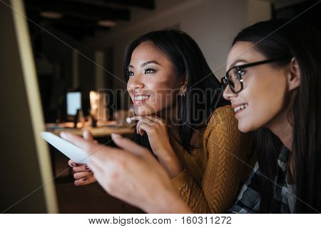 Happy businesswomen working late at night in office with computer. Looking at computer and pointing on it..