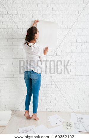 Back of woman looking at empty paper on wall mockup. Artist holding white whatman against white brick side of workshop, free space for text or advertisement