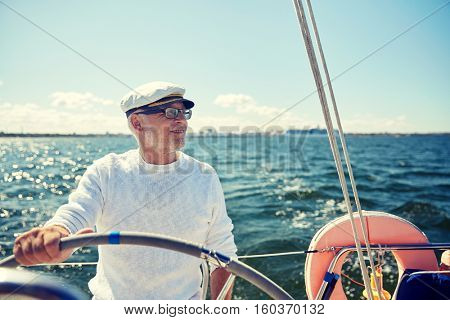 sailing, age, tourism, travel and people concept - happy senior man in captain hat on steering wheel and navigating sail boat or yacht floating in sea