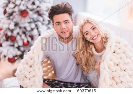 Keeping warmth. Happy nice delighted couple sitting together and covering themselves with a warm knitted plaid while having a great time