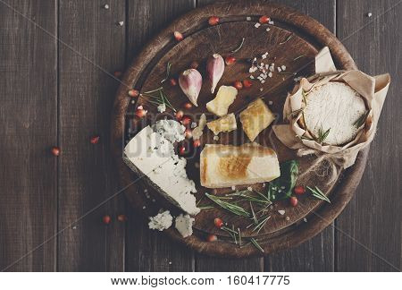 Cheese delikatessen top view on rustic wood. Wooden round desk with blue cheese roquefort, parmesan, camembert and brie cuts decorated with garlic, pomegranate and basil, french cuisine ingredients