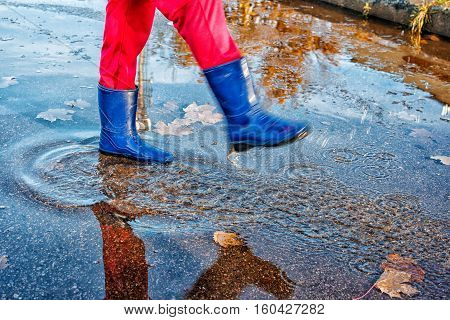 girl in blue boots and red pants standing in a puddle of water splashes with fallen yellow leaves on autuman day closeup