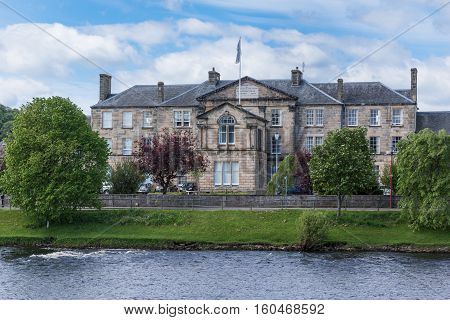 Inverness Scotland - June 1 2012: Main brown stone building of the University of Highlands and Islands seen from across the Ness River. Flag on top. Green trees. Blue sky with white clouds.